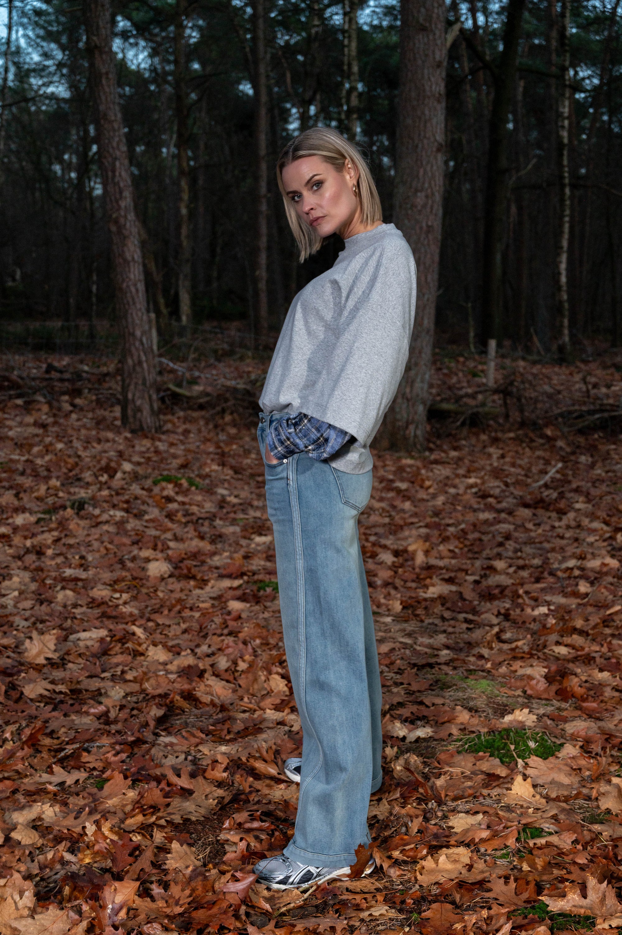 zijaanzicht van model in het bos die de nieuwe herfstcollectie toont met een oversized grijs shirt en stoere spijkerbroek