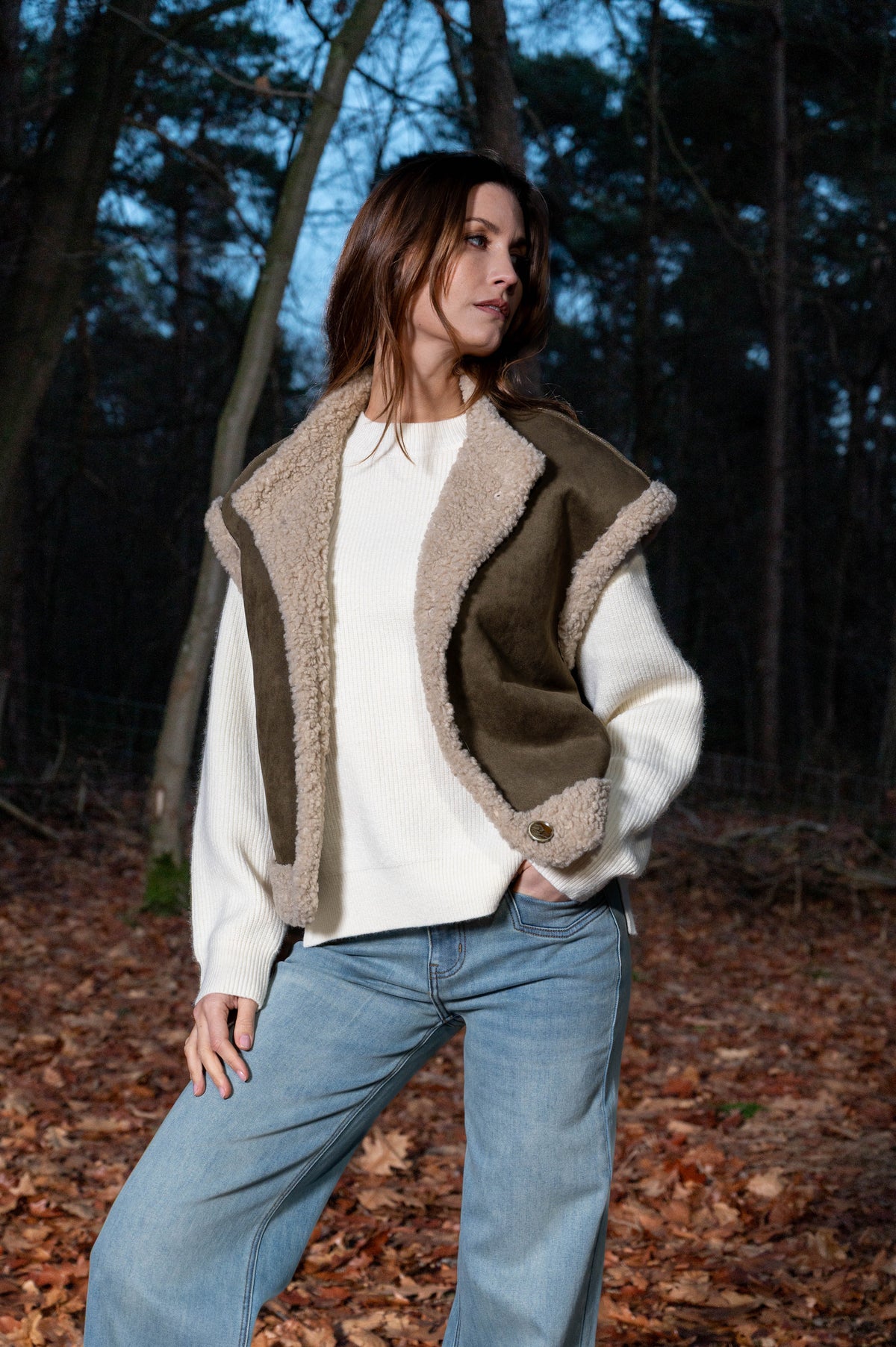 model draagt een mosgroen gilet over een offwhite trui op een lichte spijkerbroek in het bos