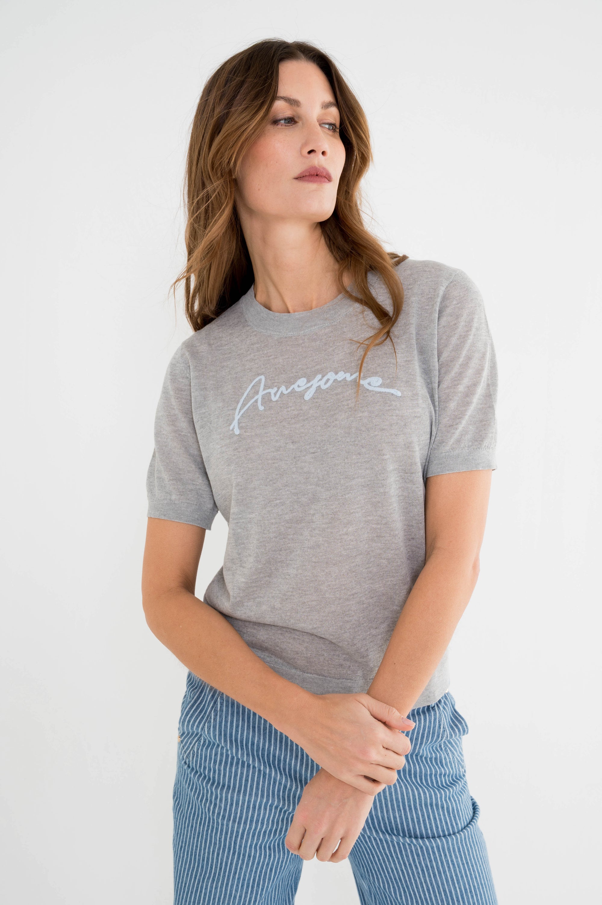 model draagt lichtgrijs t-shirt met tekst awesome in lichtblauw op de borst boven een gestreepte blauwe spijkerbroek