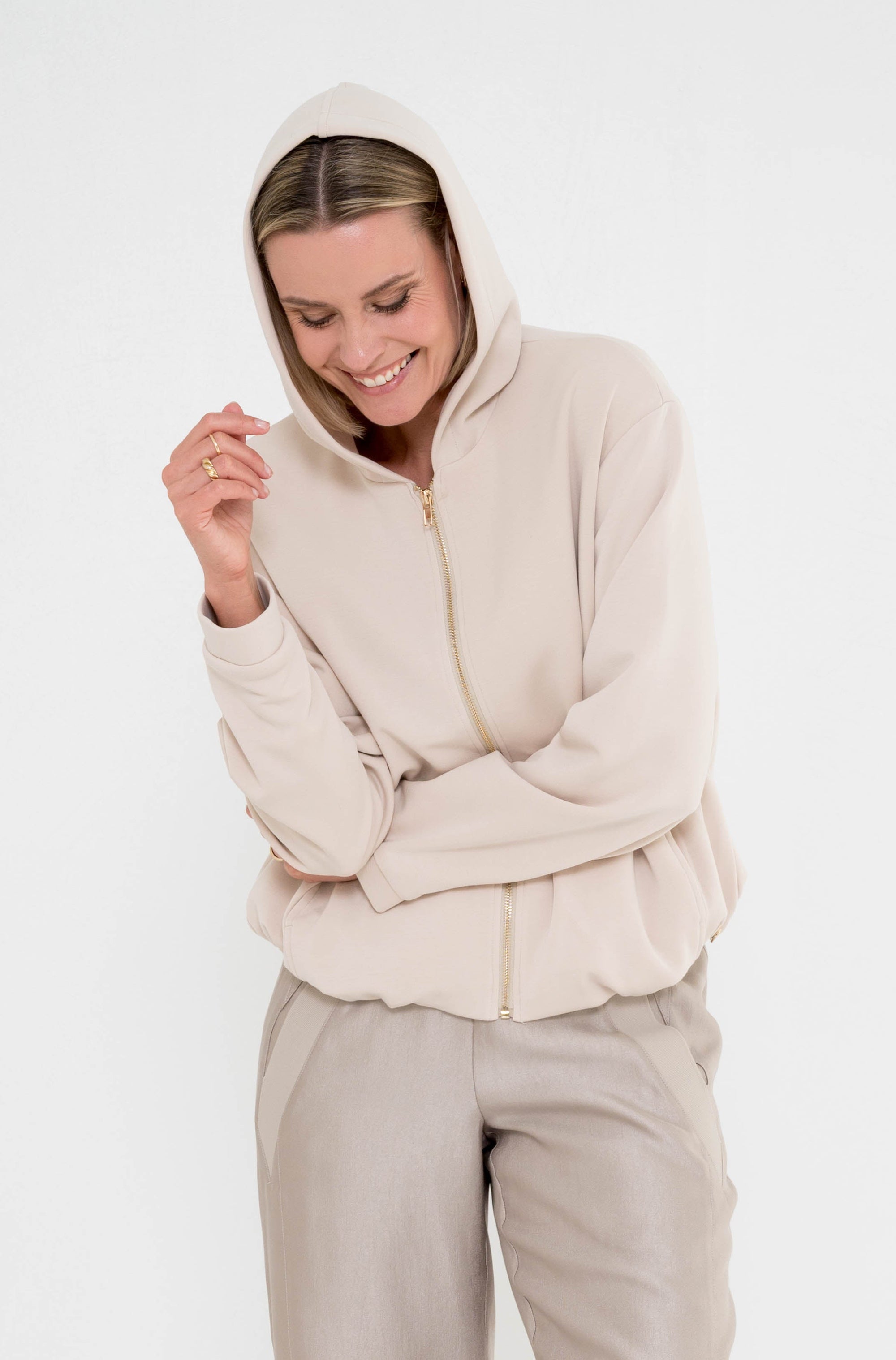 zandkleurig vest met capuchon en ritssluiting op een greige broek gedragen